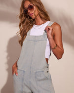 Woman wearing light wash denim overalls over white crop top, aviator sunglasses, layered gold necklaces, casual summer style