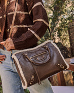 A person wearing a brown knit cardigan and light blue jeans carries a brown leather shoulder bag with white shearling-style trim and gold-tone hardware in an outdoor setting.