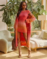 A woman stands in a bright, sunlit living room wearing a fuzzy knit mini dress with a warm gradient from coral pink to orange and red, styled with a matching long cardigan, tan knee-high boots, and a brown tote bag.