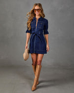 A woman models a dark blue denim shirt dress with a front zipper, ruffled sleeves, and a matching tie belt. She is wearing tan knee-high leather boots and holding a woven tan clutch against a gray studio backdrop.