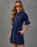 A woman modeling a dark blue denim mini dress with puff sleeves, a front zipper, chest pocket, and self-tie belt, posed against a textured gray studio background.