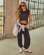 Woman in charcoal crop top, wide-leg sweatpants, Adidas Sambas, quilted tote bag, gray hoodie tied at waist, industrial backdrop