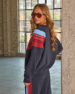 Woman wearing navy colorblock sweatsuit with red and blue stripe details, posing in industrial loft, red sunglasses, wavy blonde hair