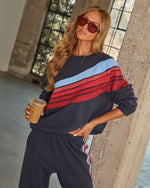 Woman in navy striped colorblock sweatsuit holding iced coffee, wearing red tinted sunglasses, leaning against concrete pillar