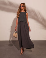 Woman in charcoal knit top layered over flowing maxi dress, holding woven bag, wearing strappy sandals and sunglasses