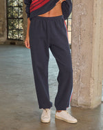 Navy blue jogger sweatpants with red side stripe, elastic waist and cuffs, styled with white sneakers on female model