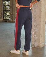 Navy blue jogger sweatpants with red and light blue side stripes, worn with white gold sneakers, back view