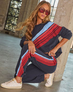 Woman wearing navy tracksuit with red and blue diagonal stripes, white sneakers, and red sunglasses, posed in industrial setting