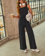 Woman wearing black wide-leg jumpsuit with square neckline, white headphones, silver sneakers, holding iced coffee outdoors