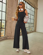 Woman in black wide-leg jumpsuit holding iced coffee, wearing white headphones and silver sneakers in industrial loft setting