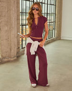 Woman wearing burgundy wide-leg pants and matching crop henley top with gray hoodie tied at waist, sunglasses, white sneakers