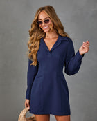 Navy blue long-sleeve polo mini dress with V-neckline and pockets, styled with cat-eye sunglasses and woven bag