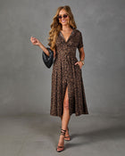 A woman models a brown and black leopard-print midi dress with short sleeves, a V-neckline, and a front slit. She is wearing black strappy heels, sunglasses, and holding a black textured clutch against a gray studio backdrop.