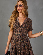 A woman modeling a brown and black leopard print dress with a V-neck, short sleeves, and a matching belt with a gold buckle, set against a solid gray background.