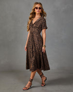 A woman models a brown and black leopard-print midi dress with short sleeves, a V-neckline, and a tie waist, styled with sunglasses, bracelets, and strappy high-heeled sandals against a gray studio backdrop.