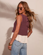 Woman wearing mauve knit sweater vest with cream ruffle hem, lace-up back detail, paired with light wash jeans and gold accessories