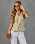 Woman wearing sage green sleeveless drawstring peplum blouse, white jeans, tan crossbody bag, and gold-frame sunglasses