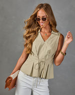 Woman wearing sage green sleeveless drawstring peplum blouse, white jeans, gold accessories, and oval sunglasses
