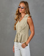 Woman wearing sage green sleeveless drawstring peplum blouse with collar, white pants, gold bangles, and oval sunglasses