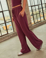 Burgundy wide-leg drawstring sweatpants with elastic waist, paired with matching crop top and white sneakers, industrial loft setting