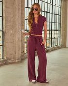 Woman wearing burgundy matching set: crop henley top and wide-leg drawstring pants, styled with sunglasses, in industrial loft setting