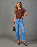 Woman wearing brown ringer tee, light wash straight-leg jeans with seam detail, black strappy heels, holding dark woven bag