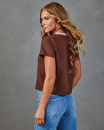 Woman wearing brown cropped t-shirt with pink collar and blue jeans, posed from behind showing back view of casual outfit