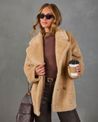 A woman models a camel-colored teddy coat with a double-breasted button closure, styled with a brown top, dark brown leather pants, sunglasses, and a brown quilted handbag against a textured gray studio background.