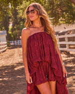 Woman in burgundy strapless tiered ruffle romper dress with drawstring waist, gold jewelry, aviator sunglasses, outdoor setting