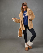A woman models a beige faux-shearling coat over a blue sweatshirt, paired with black leggings, brown suede boots with white shearling trim, a brown cap, sunglasses, and a quilted tan handbag, set against a gray studio backdrop.