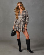 A woman models a beige and black plaid mini shirt dress with a tie waist, paired with black knee-high boots and holding a black textured handbag against a gray studio backdrop.