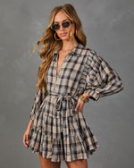 A woman modeling a beige, brown, and black plaid mini dress with long balloon sleeves, a tie waist, and a ruffled hem, set against a textured gray studio background.