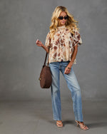 Woman wearing cream floral flutter-sleeve blouse, light wash straight-leg jeans, nude strappy heels, carrying brown suede shoulder bag