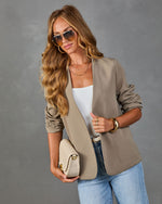 Woman wearing taupe open-front blazer, white tank, jeans, gold accessories, and sunglasses, holding beige woven clutch bag