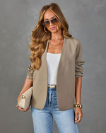 Woman wearing taupe open-front blazer, white tank, blue jeans, gold accessories, and tinted sunglasses holding cream clutch