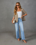 Woman wearing khaki open-front blazer, white bodysuit, light-wash wide-leg jeans, nude strappy heels, and cream mini bag