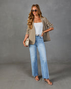 Woman wearing khaki open-front blazer, white bodysuit, light-wash wide-leg jeans, nude strappy heels, and cream mini bag