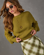 Woman in olive green ribbed knit sweater with cable sleeves, plaid midi skirt, oval sunglasses, gold hoop earrings and pendant necklace