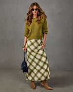 Woman wearing an olive green knit sweater, plaid satin midi skirt, tan suede heeled boots, gold bangles and sunglasses
