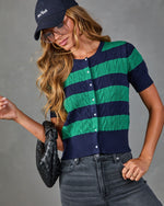 Woman wearing navy and green striped cable-knit cardigan, dark jeans, New York cap, sunglasses, holding black woven clutch