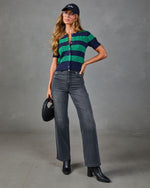 Woman wearing navy green striped cable-knit cardigan, gray wide-leg jeans, black ankle boots, baseball cap, holding woven bag