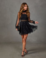 Black lace long-sleeve mini dress with sheer sleeves and high neck, styled with black strappy heels and a gold-clasp clutch