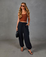 Woman wearing rust ribbed crop top, black drawstring balloon pants with ankle ties, and black strappy heels, holding quilted bag