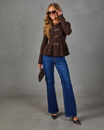 A woman modeling a dark brown long-sleeve peplum top with decorative gold-toned buttons, paired with blue flare jeans, dark pointed heels, and sunglasses, standing against a gray studio backdrop.