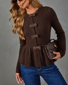 A woman wearing a brown knit sweater with decorative jeweled bow details down the front, paired with blue jeans and holding a small brown handbag against a plain gray studio background.