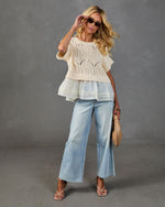 Woman wearing cream crochet knit top with ruffled hem, wide-leg light wash jeans, nude heels, holding woven clutch