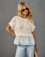 Woman wearing cream cable-knit crop sweater with chiffon peplum hem over light wash jeans, styled with tortoise sunglasses