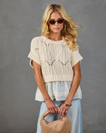 Woman wearing cream pointelle knit crop sweater top layered over white blouse, light wash jeans, woven tan bag, retro sunglasses