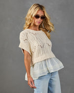 Woman wearing cream pointelle knit short-sleeve sweater with chiffon ruffle hem, light wash jeans, and tortoise sunglasses