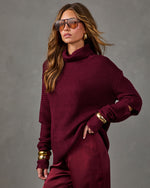 Woman in a burgundy chunky knit turtleneck sweater and satin pants with gold bangles, aviator sunglasses and gold earrings